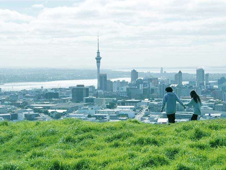 The Auckland city skyline and the Sky Tower seen from a grassy hilltop, with two people walking in the foreground