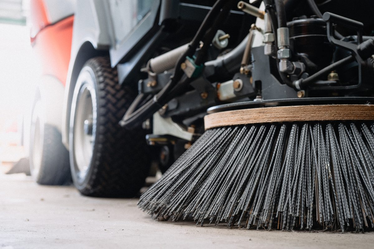 A close-up of the rotating side brush and front wheel of a street sweeper on concrete