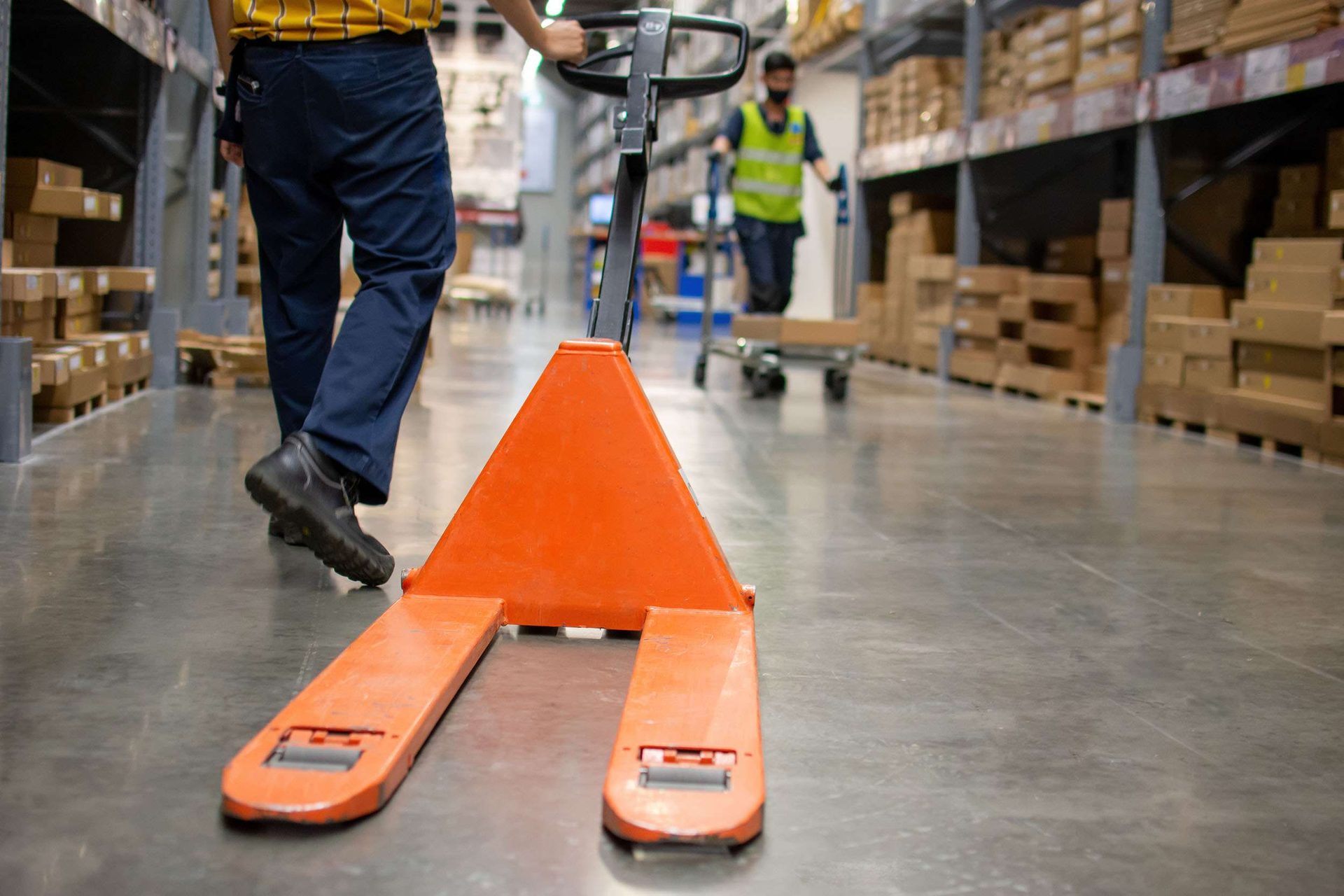 A worker pulling an orange hand pallet truck along a polished warehouse aisle, with another worker in a high-visibility vest behind