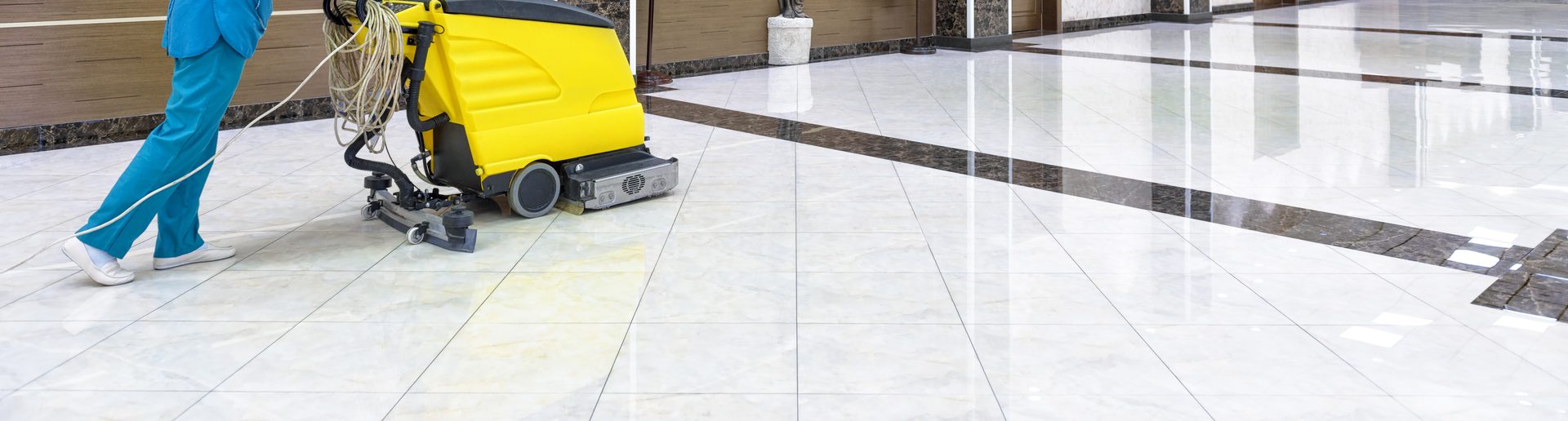 A cleaner in blue overalls guiding a yellow walk-behind floor scrubber across a tiled floor