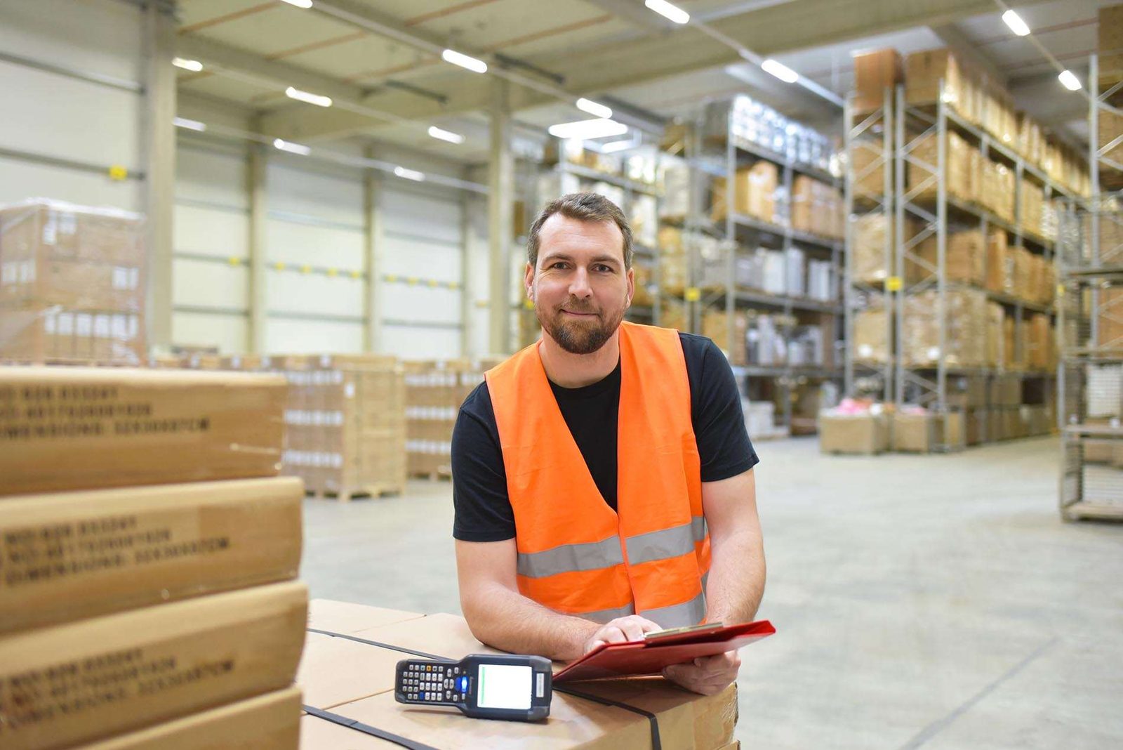 A warehouse worker in an orange high-visibility vest leaning on a stack of cartons with a clipboard and a handheld scanner, racking behind