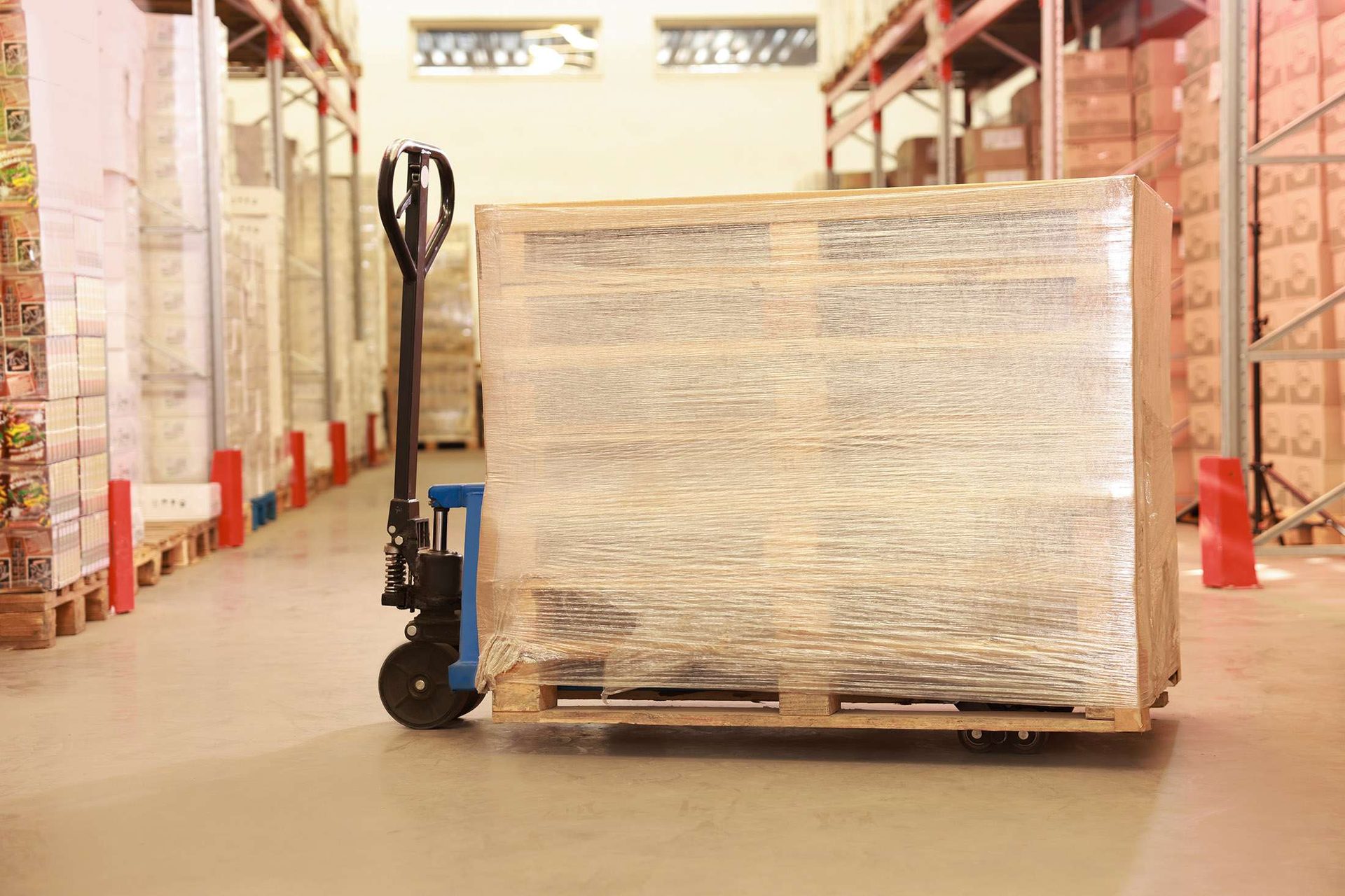 A pallet of goods wrapped in stretch film sitting on a blue hand pallet jack in a warehouse aisle